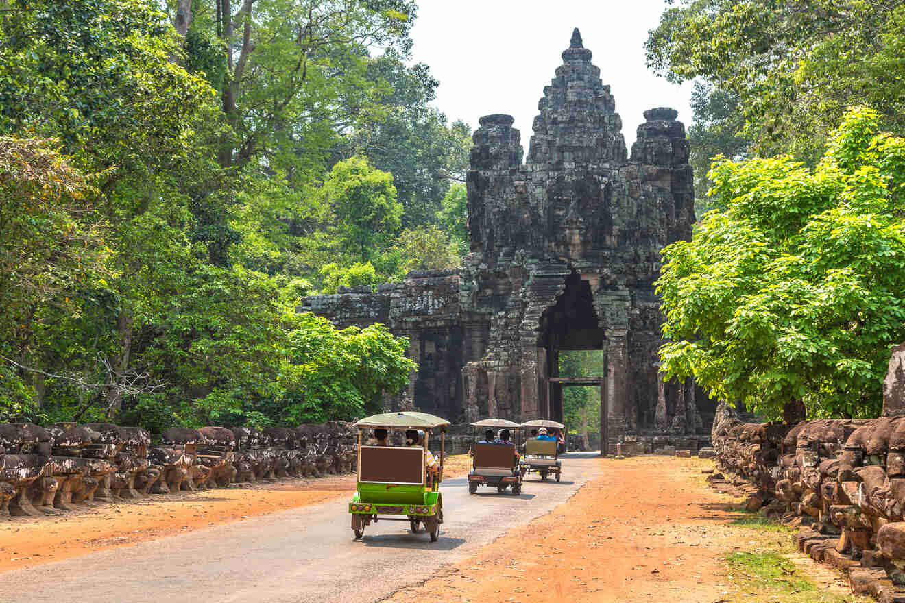 Traditional Khmer tuk-tuks driving on a dirt road leading to an ancient temple gate, flanked by stone sculptures, amidst lush tropical greenery